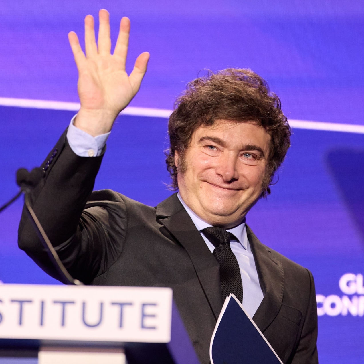 Javier Milei, a white middle-aged man with dark brown hair, smiles and waves with his right hand. He wears a dark suit and tie. The background is mauve and purple.